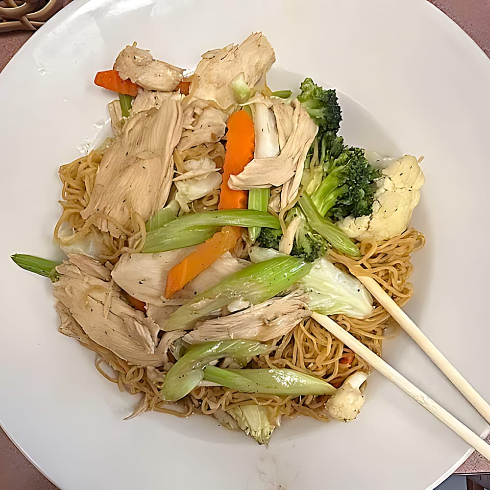 Chicken stir-fried noodles with broccoli, carrots, celery, and cauliflower, served with chopsticks.