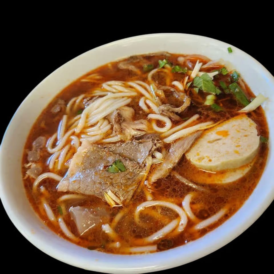 A close-up of a bowl of Vietnamese pho with noodles, beef, and a slice of sausage.